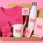 Pack de productos promocionales personalizados con logotipo de Ozé Creative Studio, incluyendo camiseta, calcetines, taza, libreta, bolígrafo, pegatinas y botella, sobre fondo rosa.