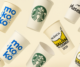 Vasos reutilizables personalizados con distintos logotipos de marcas y eventos, flotando sobre fondo beige