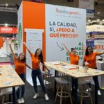 Cuatro miembros del equipo de Realisaprint.es con camisetas naranjas posan sonrientes y con los brazos abiertos en su stand de feria, decorado con mesas altas de madera y un cartel con el eslogan “La calidad, sí, ¿pero a qué precio?”.