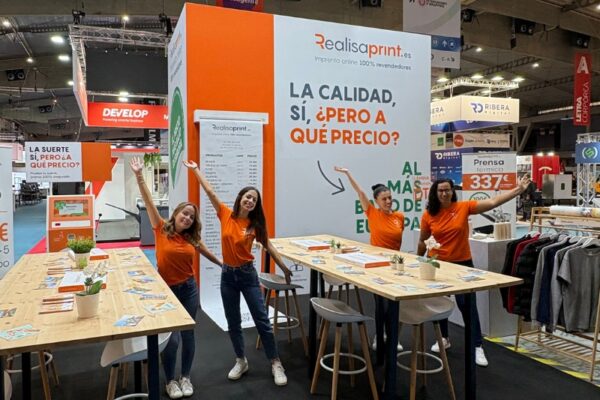 Cuatro miembros del equipo de Realisaprint.es con camisetas naranjas posan sonrientes y con los brazos abiertos en su stand de feria, decorado con mesas altas de madera y un cartel con el eslogan “La calidad, sí, ¿pero a qué precio?”.