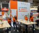 Cuatro miembros del equipo de Realisaprint.es con camisetas naranjas posan sonrientes y con los brazos abiertos en su stand de feria, decorado con mesas altas de madera y un cartel con el eslogan “La calidad, sí, ¿pero a qué precio?”.