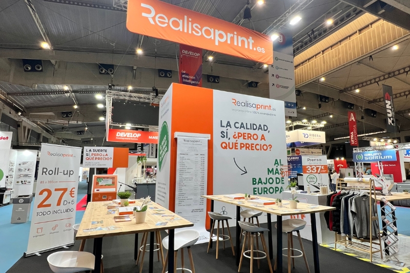 La imprenta online preferida de los profesionales Vista del stand de Realisaprint.es en una feria gráfica, con mesas altas, folletos, un cartel que indica precios y el eslogan “La calidad, sí, ¿pero a qué precio?”, junto a una gran pancarta colgante de la marca.