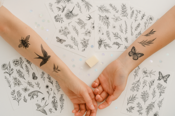 Dos brazos con tatuajes temporales en blanco y negro de flores, mariposas y aves, junto a varias láminas con diseños botánicos.